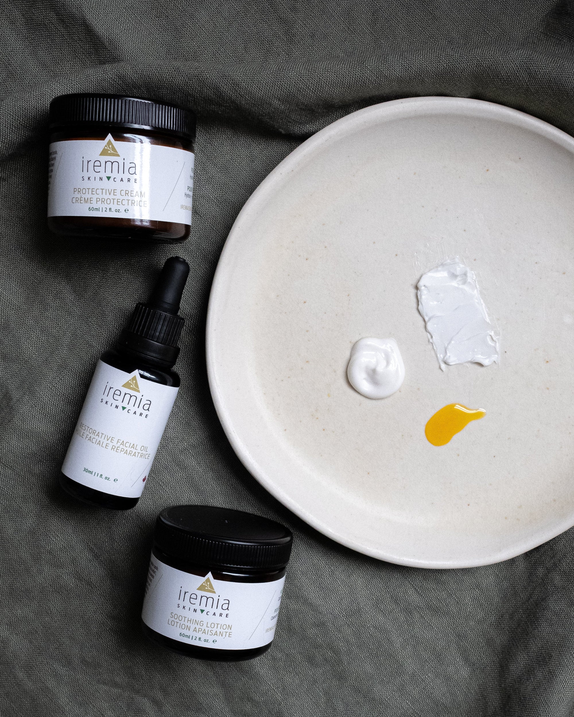 Iremia Skincare's Soothing Lotion, Restorative Facial Oil and Protective Cream are position on the left of the image next to a neutral plate which shows the textures of the products. 